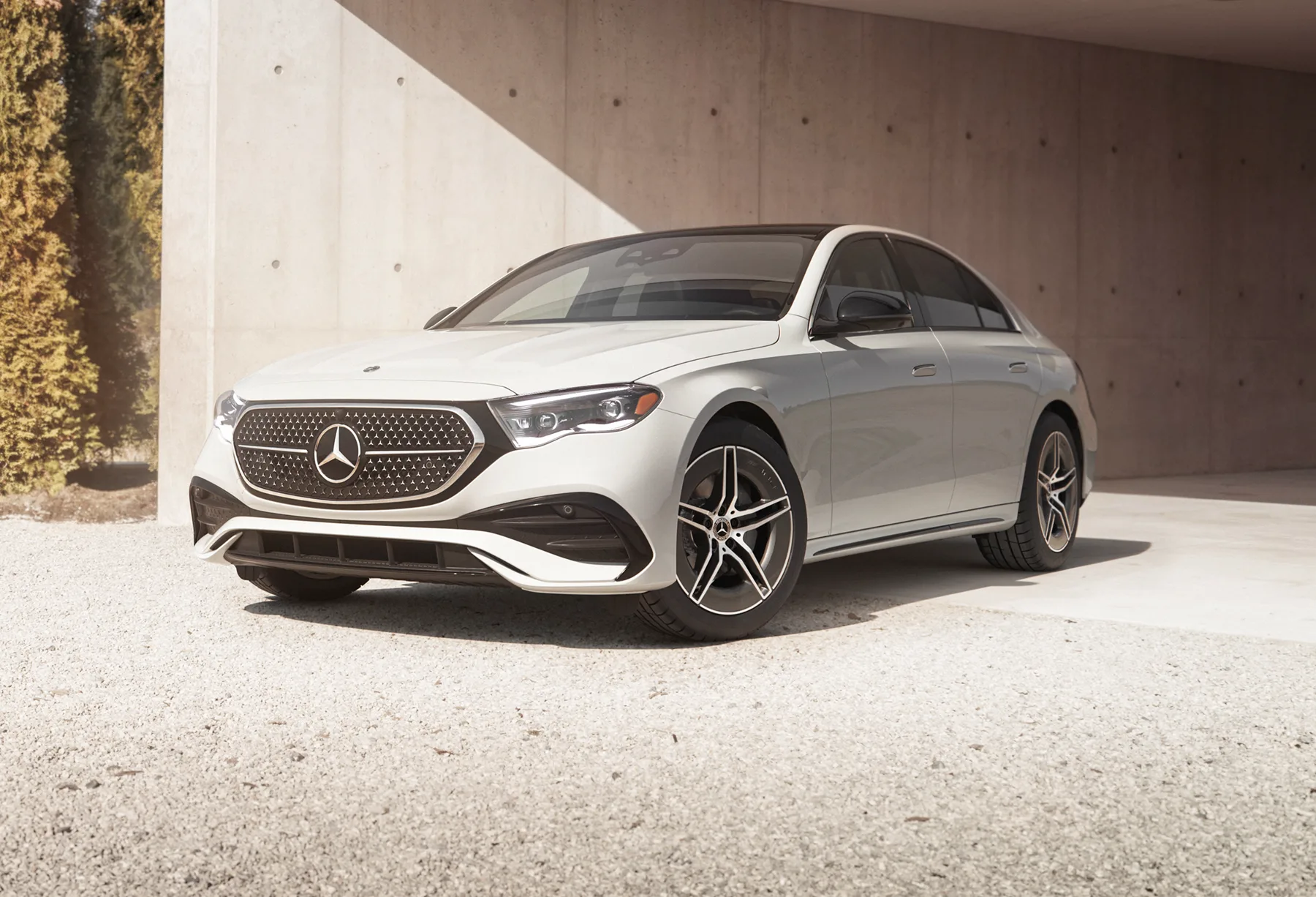 Silver 2026 Mercedes-Benz E-Class sedan, front three-quarter on gravel under a concrete overhang