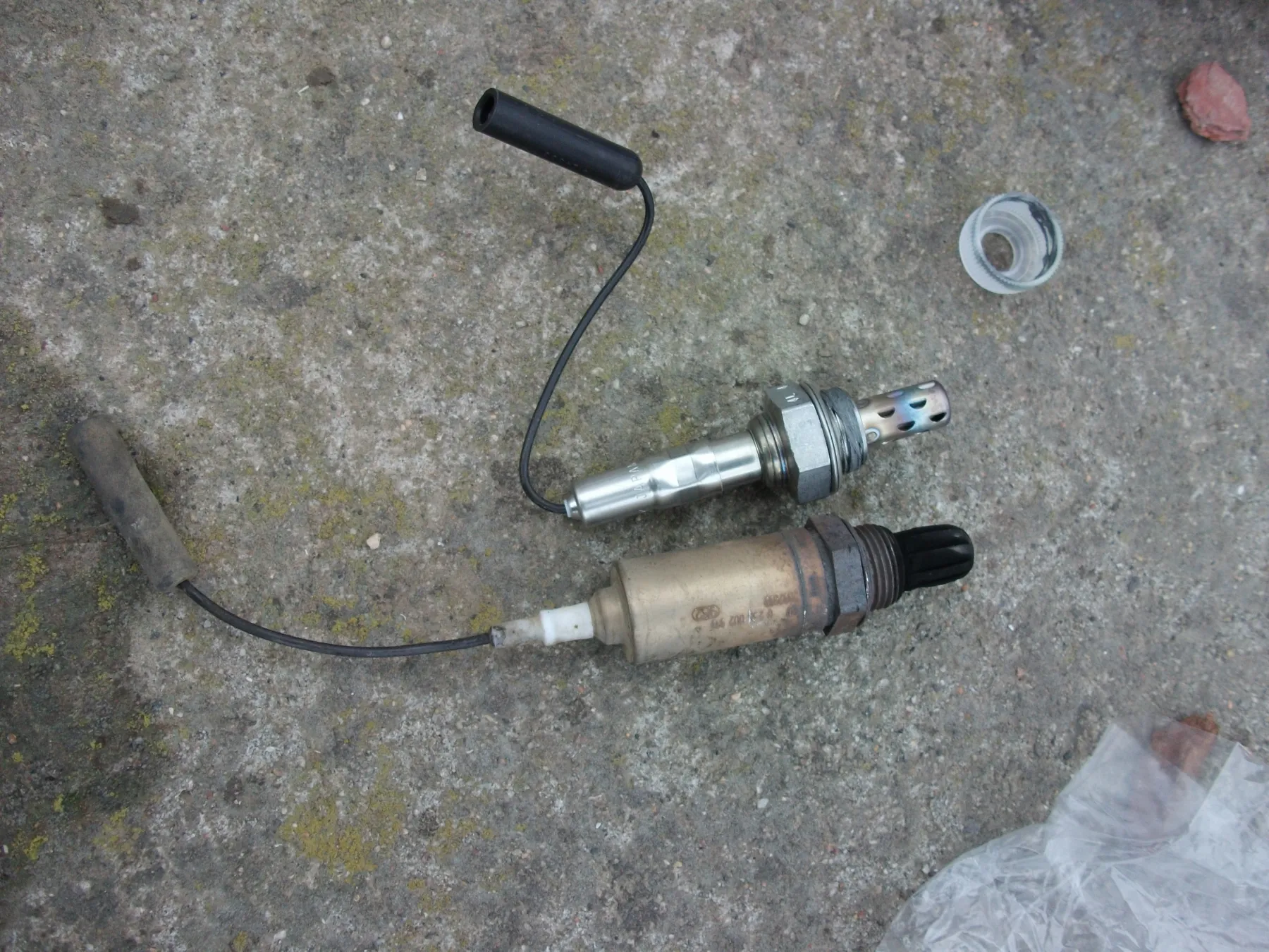 New oxygen sensor above a soot-black used sensor on concrete